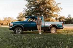 Person leaning against a teal Ford pickup truck parked on green grass near trees at golden hour.