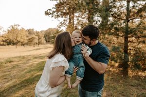 A happy young family with a laughing baby boy being kissed by both parents outdoors in a sunny autumn park.