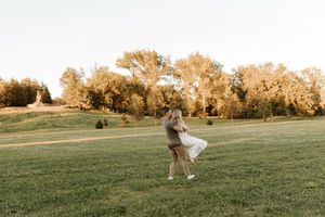 Couple dancing joyfully in a sunlit green field surrounded by golden trees during a romantic outdoor photo session.