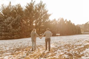 Couple holding hands in snowy field with sunlit pine trees in background during golden hour.