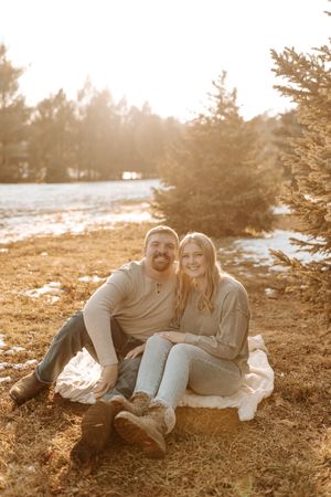A smiling couple sits together on a blanket outdoors near pine trees with golden sunlight in a winter setting.