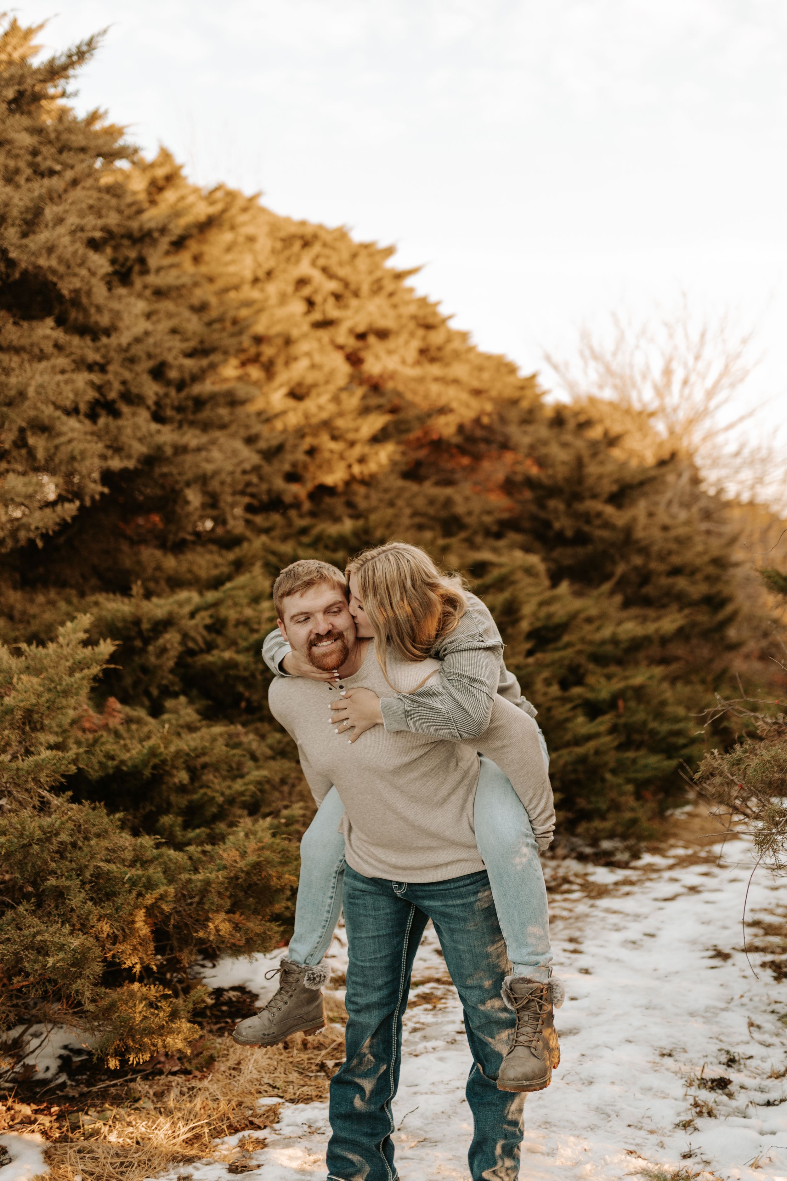 A bearded man gives a woman a piggyback ride through a snowy tree farm path at golden hour.