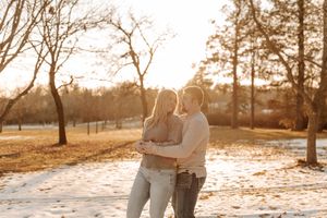 Couple embracing in a snowy park at golden hour, surrounded by bare trees in warm winter sunlight.