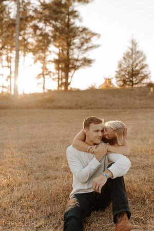 Couple embracing in golden field at sunset, woman hugging man from behind among dry grass with trees in background.
