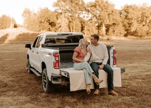 Couple sitting on tailgate of white pickup truck in golden hour field, sharing a romantic moment.