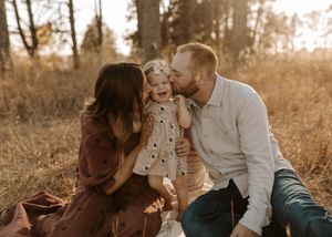 A happy family of three shares a joyful moment outdoors in autumn, with parents kissing their laughing toddler daughter.