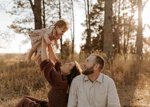Parents sit in a sunlit forest meadow as mother lifts smiling toddler in the air while father watches lovingly.