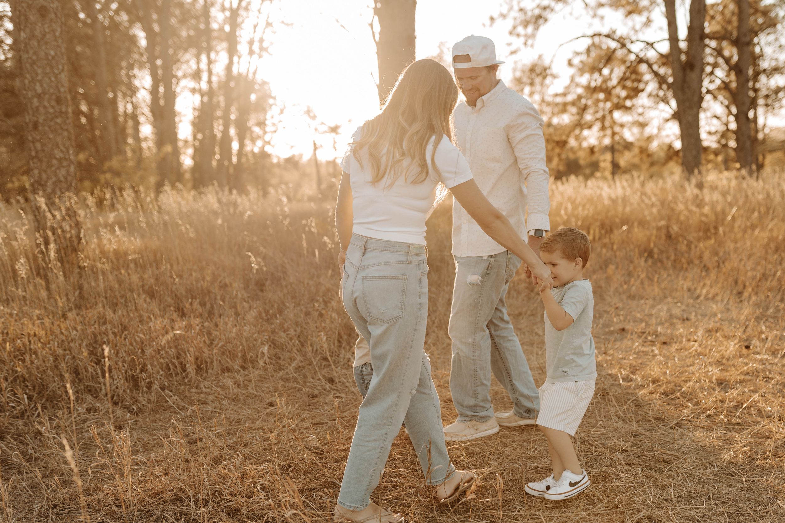 Family of three holding hands in a golden sunlit field, parents and young child enjoying a warm autumn outdoor walk.