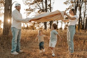 Family playing outdoors at golden hour, parents holding blanket over two young children running in a sunlit forest field.