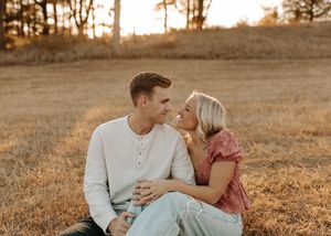 Couple sitting in golden field at sunset, gazing at each other romantically during an outdoor engagement photo session.