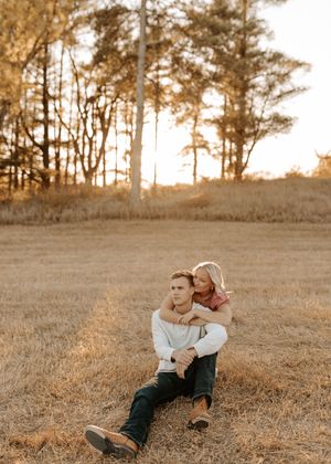 Couple sitting in golden field at sunset, man seated on grass with woman embracing him from behind, trees in background.