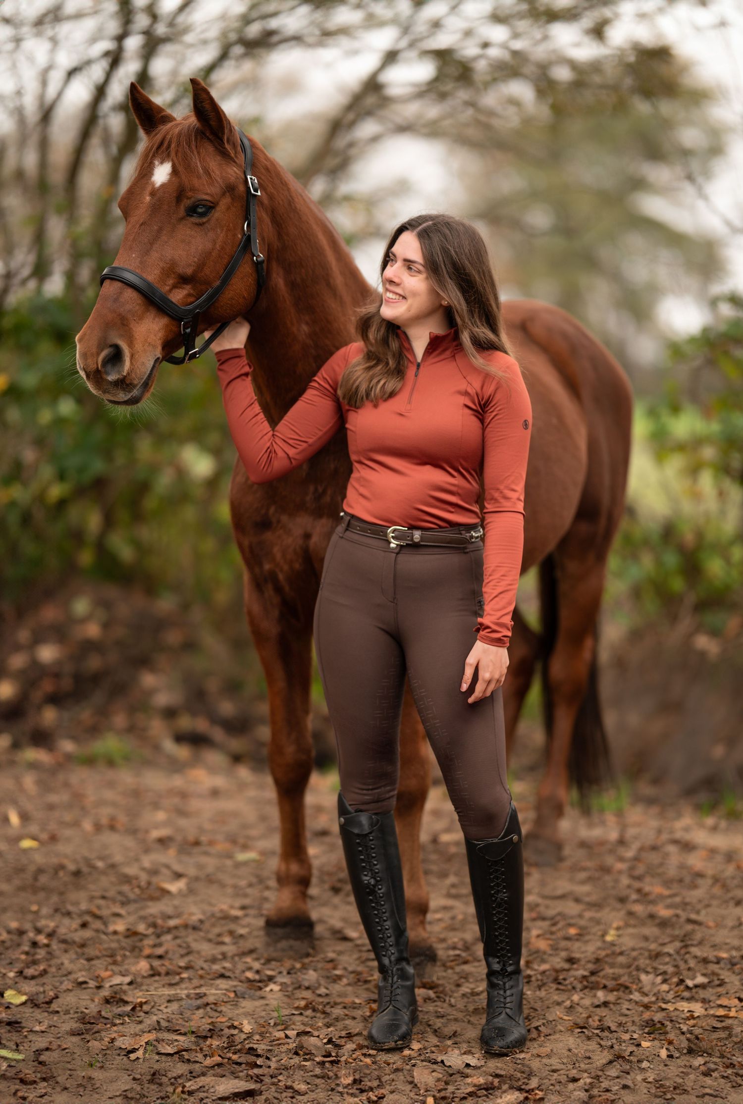 Young woman in rust top and brown riding breeches stands beside a chestnut horse outdoors.
