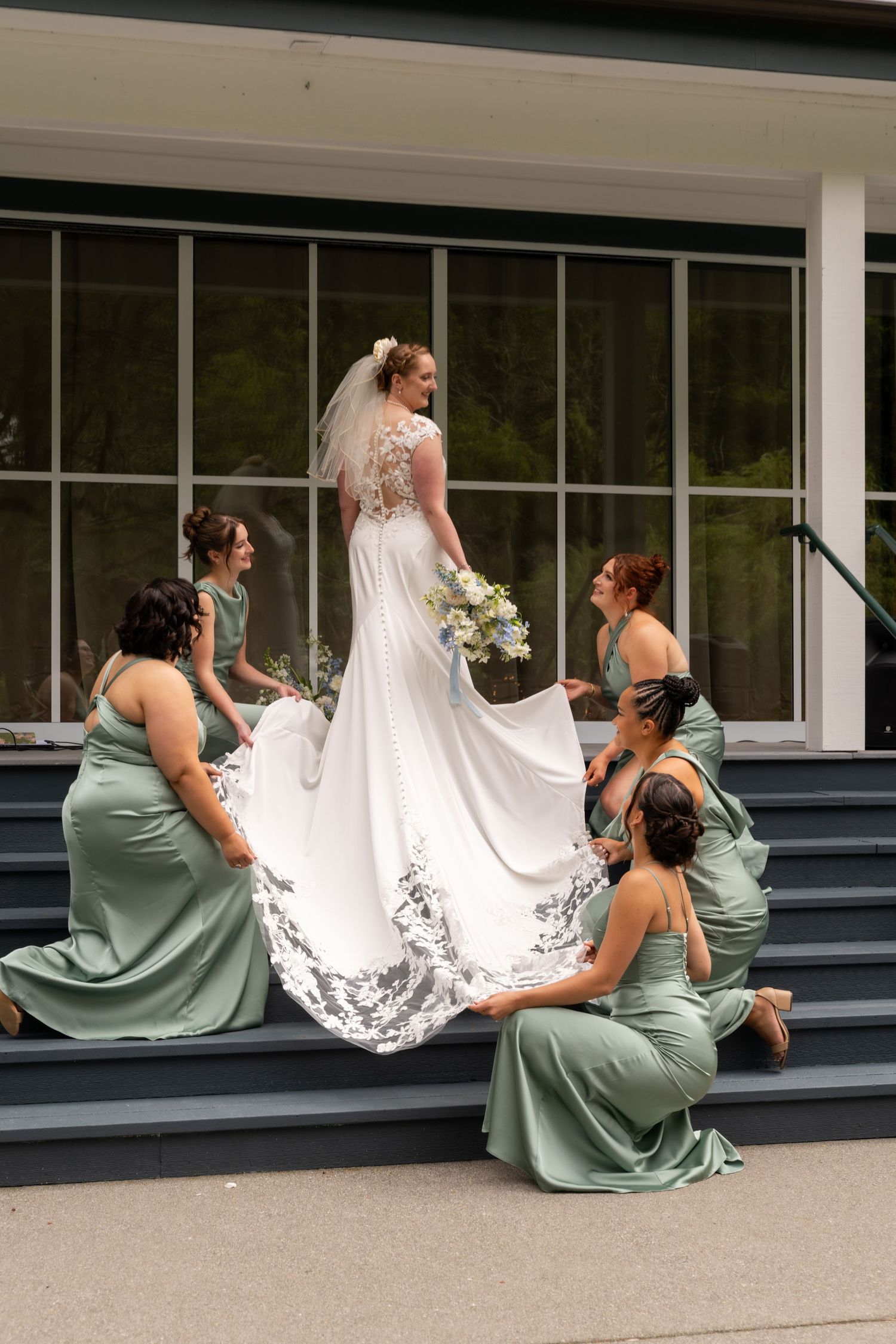 Bride in white lace gown with veil holds bouquet on steps as four bridesmaids in sage green dresses arrange her train.