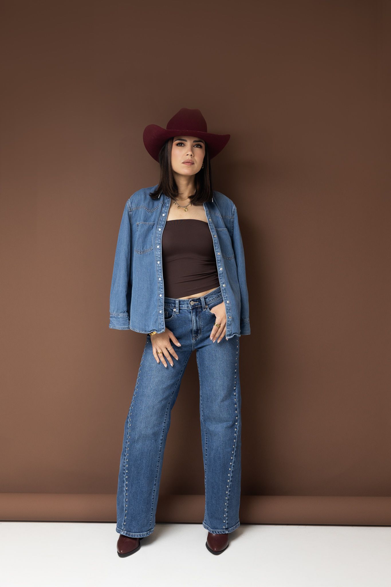 Woman in wide-leg jeans, denim shirt, brown top, and burgundy cowboy hat posing against a brown wall.