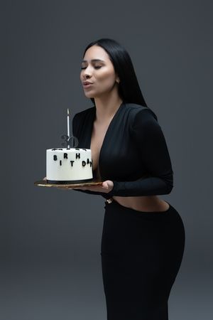 Woman in black outfit blowing out candle on white birthday cake with dark lettering on gray background.