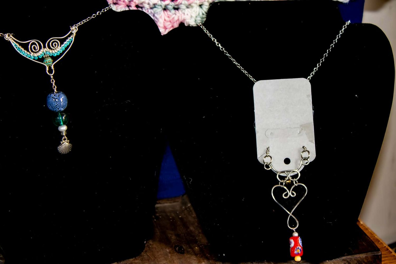 Two handmade silver necklaces on black display stands, one with blue beads and one with a heart pendant and red beads.