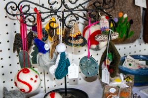 Handmade holiday ornaments displayed on a decorative metal stand at a craft fair, featuring felted and glass designs.