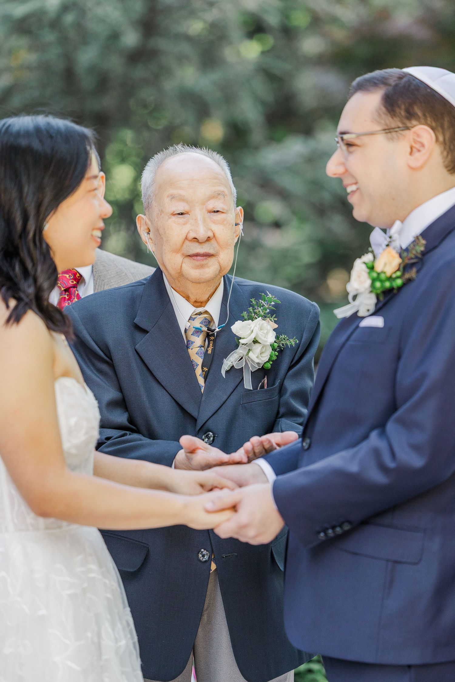 Elderly officiant joins hands of bride and groom during intimate outdoor wedding ceremony.
