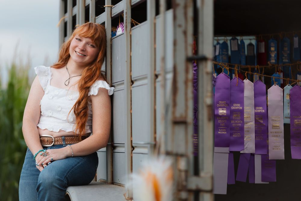 Young woman with long red hair smiles beside purple award ribbons at a county fair livestock show.