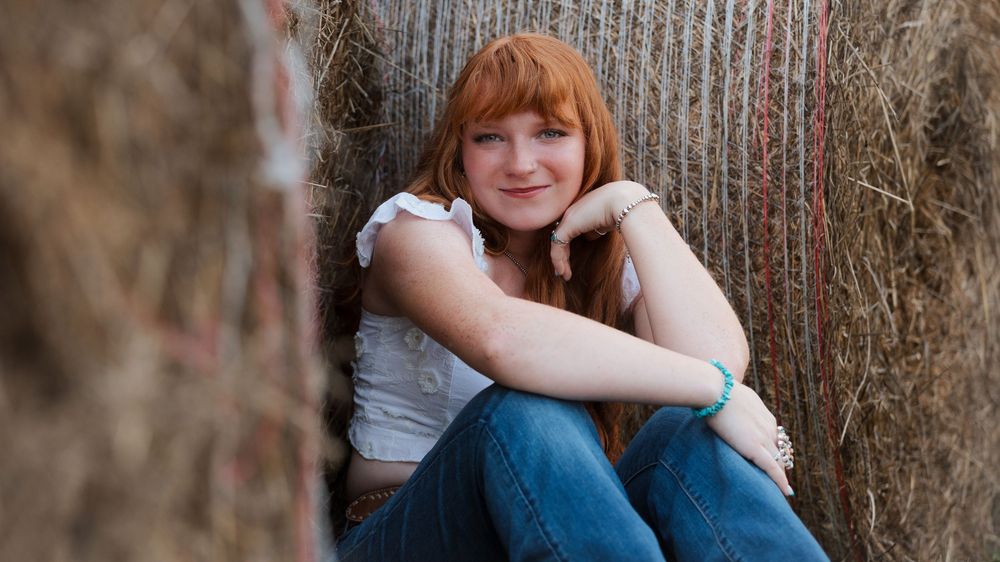 A young redhead girl sits smiling against hay bales, wearing a white top and jeans, with a turquoise bracelet.