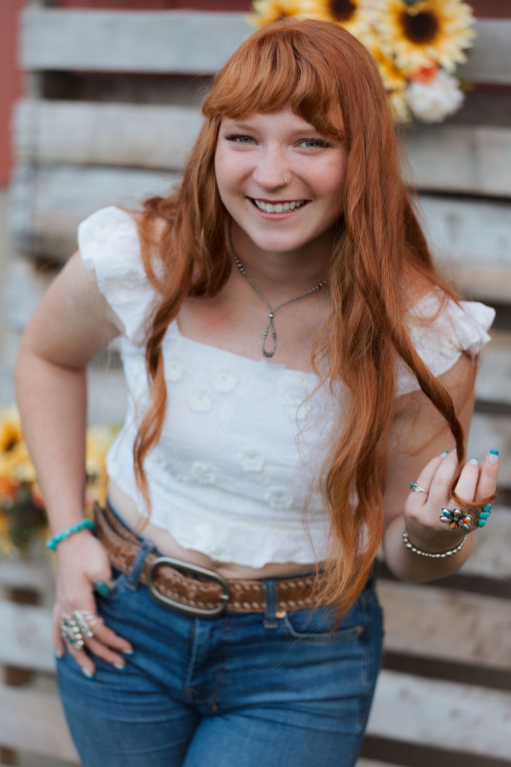 Smiling redhead teen girl in white crop top, jeans, and turquoise jewelry poses outdoors near sunflowers.