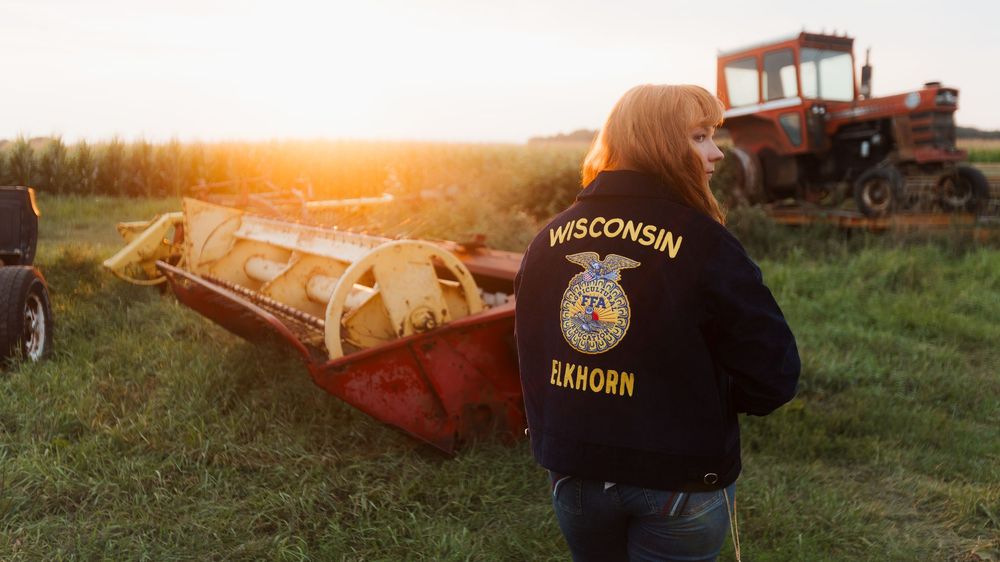 Student in Wisconsin Elkhorn FFA jacket stands beside farm equipment at sunset with red tractor in background.