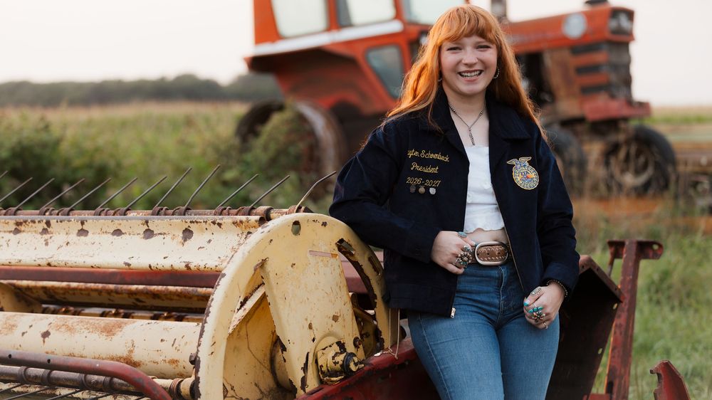 Young woman in FFA jacket smiling beside farm equipment with a red tractor in a rural field.