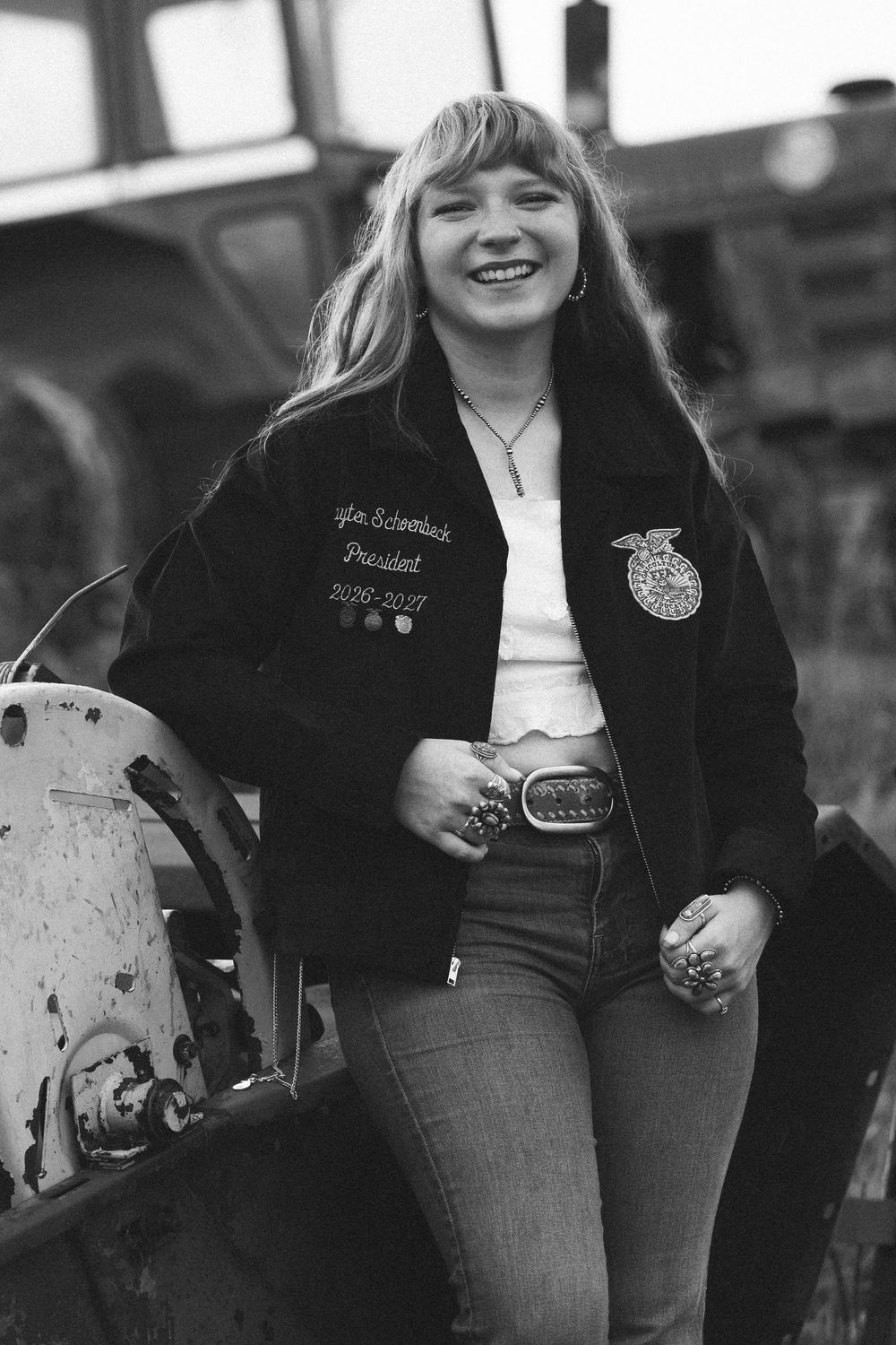 Young woman in FFA jacket smiling outdoors near farm equipment in black and white photo.