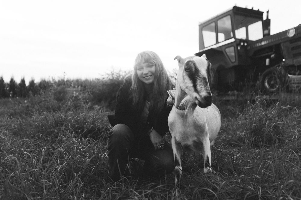 Black and white photo of a smiling woman crouching beside a goat on a farm with a tractor in the background.