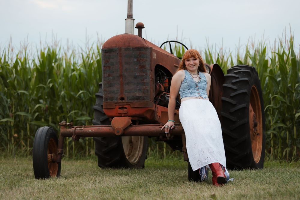 A woman in a white skirt and red boots leans against a rusty vintage red tractor in a cornfield.