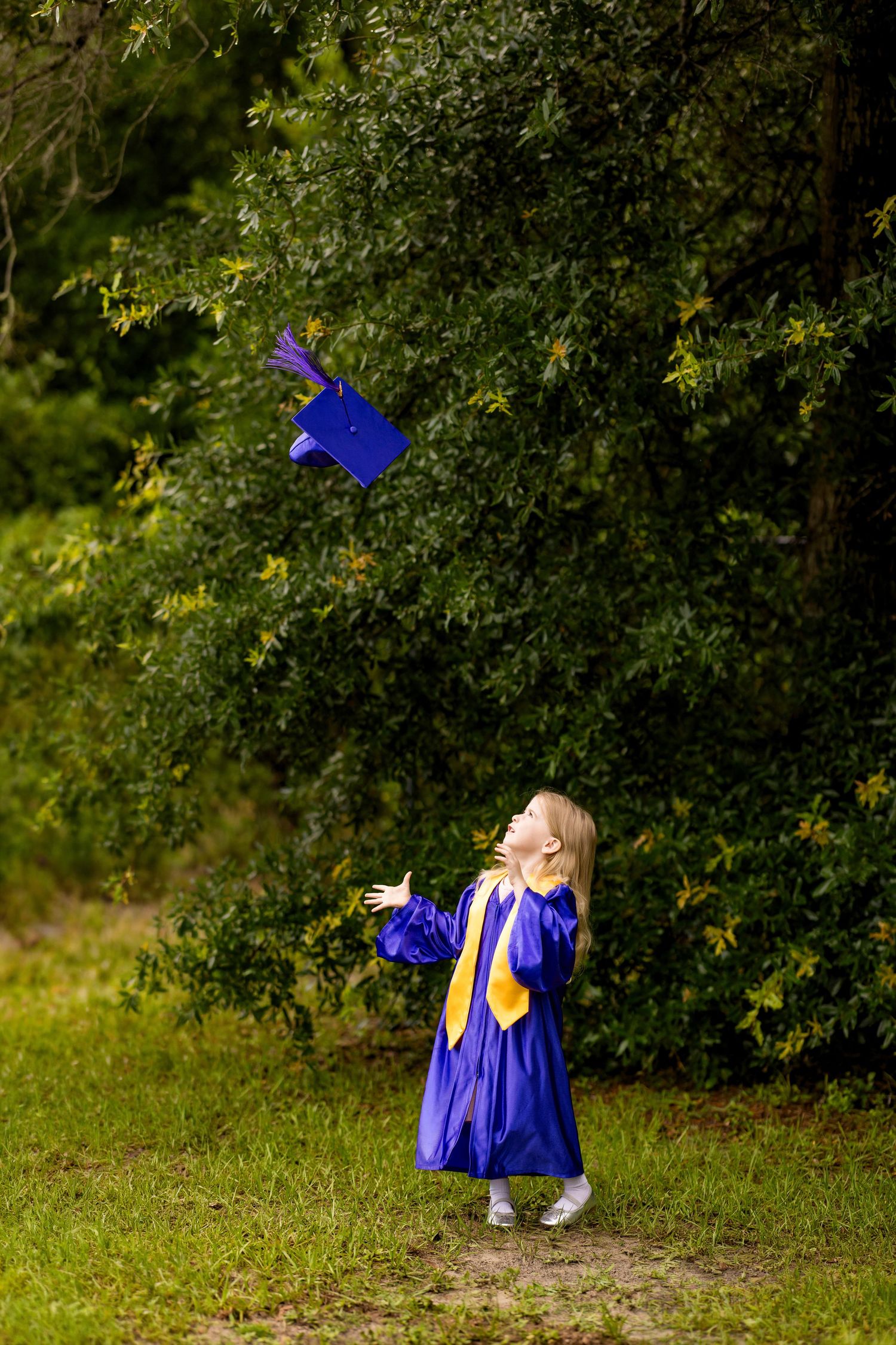 Young girl in blue graduation gown tossing cap in air outdoors, celebrating preschool or kindergarten graduation.