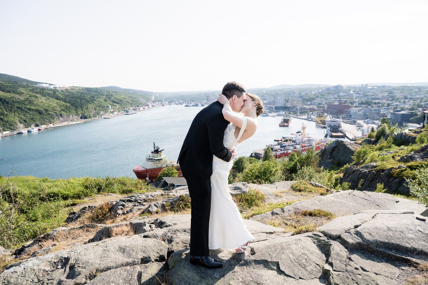 Newlywed couple cuddling each other with the stunning St. John's skyline in the background, standing on the rocks of a hill.