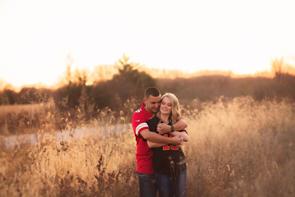Couple embracing in golden field at sunset, man in red shirt hugging woman from behind amid tall grass.