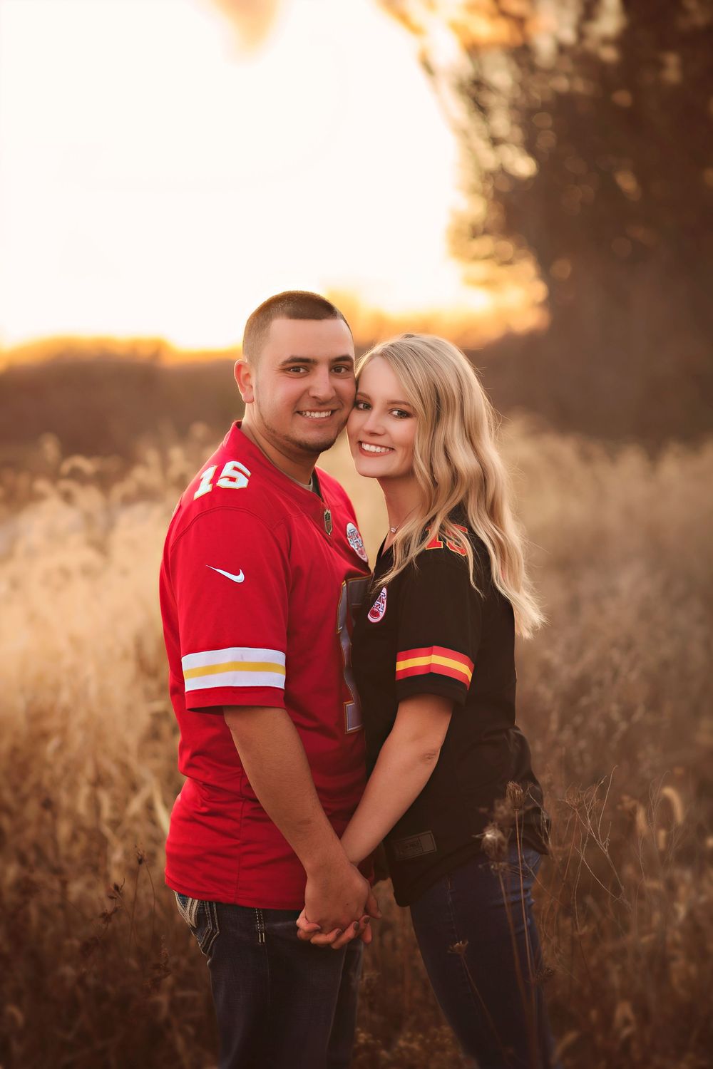 Couple in Kansas City Chiefs jerseys holding hands in a golden field at sunset.