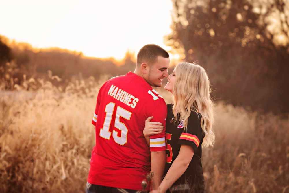 Couple sharing a romantic kiss in a golden field, wearing Kansas City Chiefs and Cleveland Browns NFL jerseys at sunset.