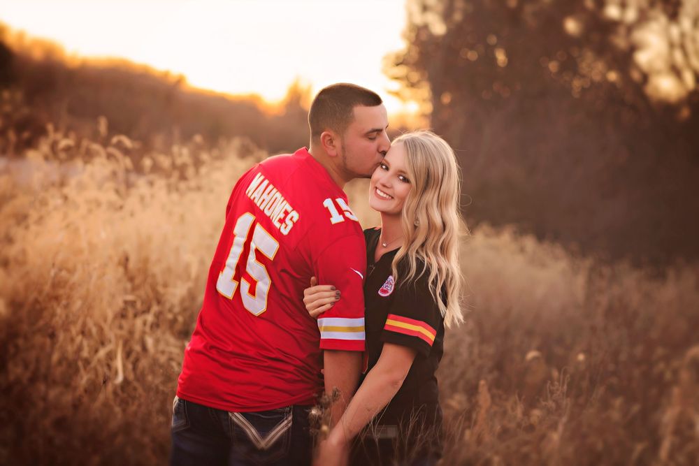 Couple in Kansas City Chiefs jerseys embracing in golden field at sunset, sharing a romantic kiss.