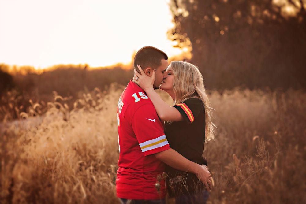 Couple kissing in golden field at sunset, man in red jersey embracing blonde woman in romantic outdoor portrait.