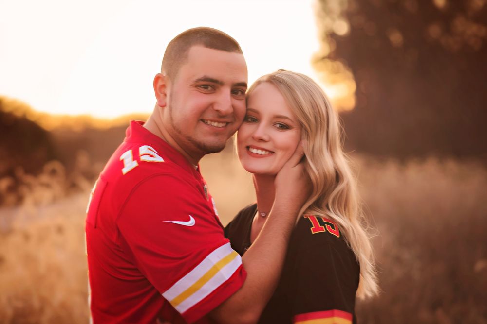 Couple posing outdoors at golden hour, man in red Kansas City Chiefs jersey, woman in black top, warm sunset background.