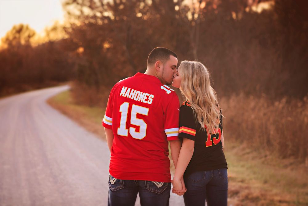 Couple in NFL jerseys kissing on a rural road at sunset, man in red Mahomes 15 Chiefs jersey, woman in Browns jersey.
