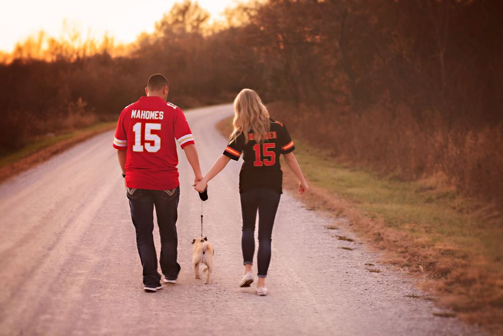 Couple in matching #15 Mahomes KC Chiefs jerseys walking dog on rural road at sunset.