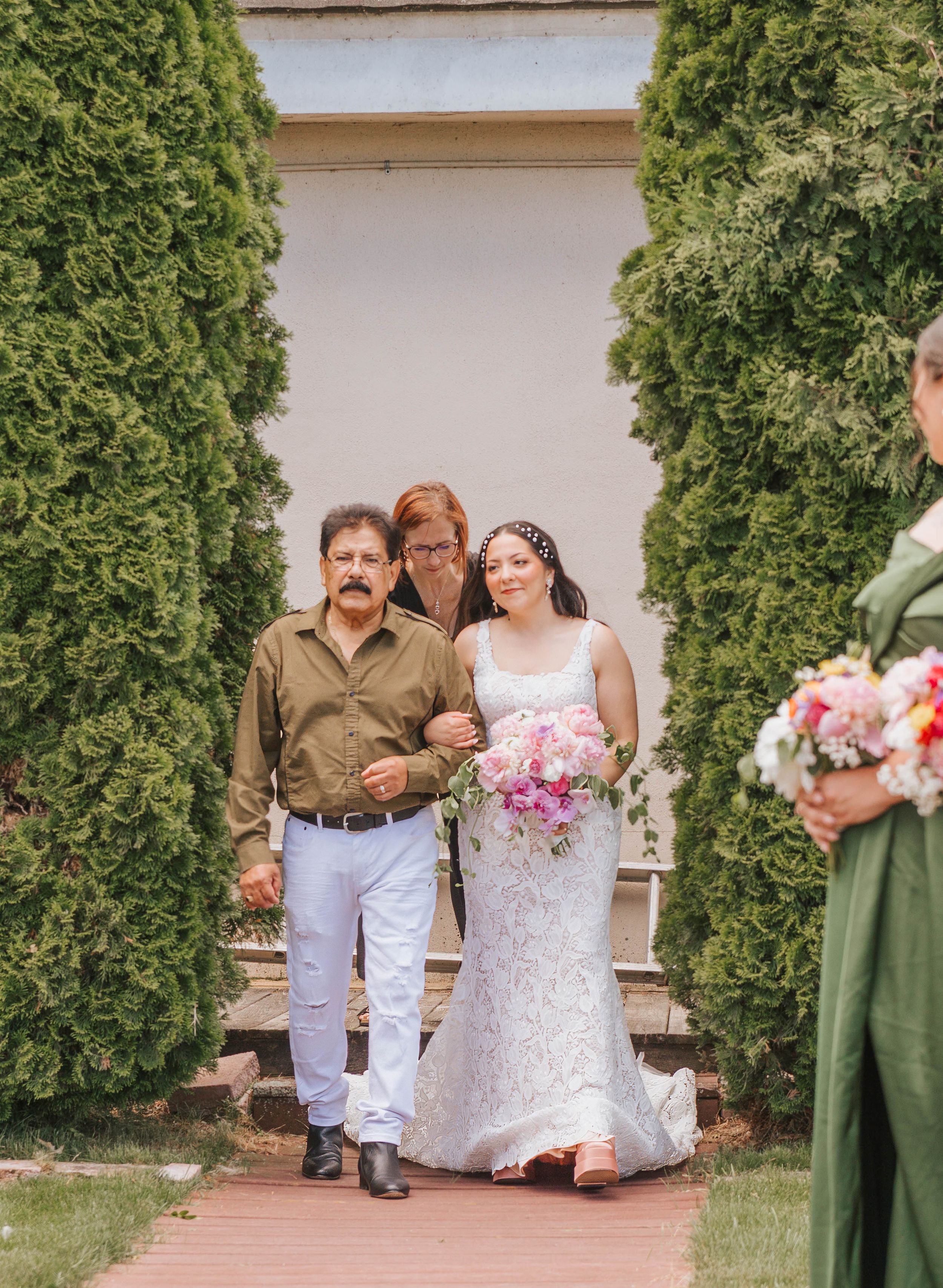 Bride in white lace gown walks with family through tree-lined garden path holding pink bouquet.