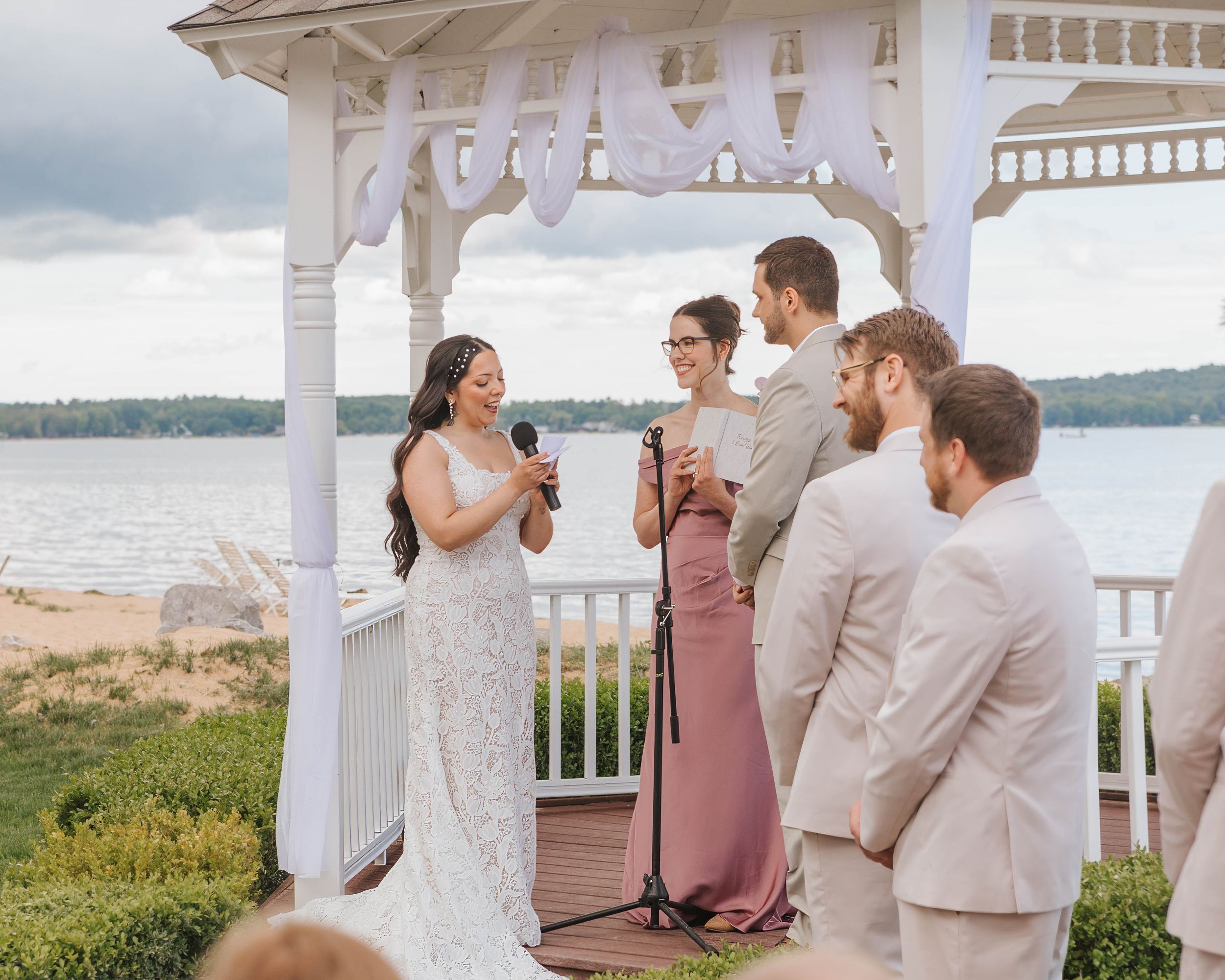 Bride reading vows at outdoor waterfront gazebo wedding ceremony with bridal party in pink and beige.