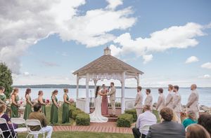 Outdoor wedding ceremony at a waterfront gazebo with bridesmaids in green dresses and guests seated on white chairs.