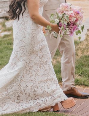 Bride in lace wedding gown holding pink floral bouquet stands beside groom in tan suit on wooden deck.