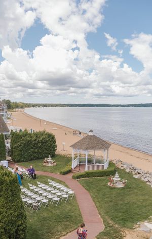 White gazebo and chairs set up for a lakeside wedding ceremony on a sandy beach under a partly cloudy sky.