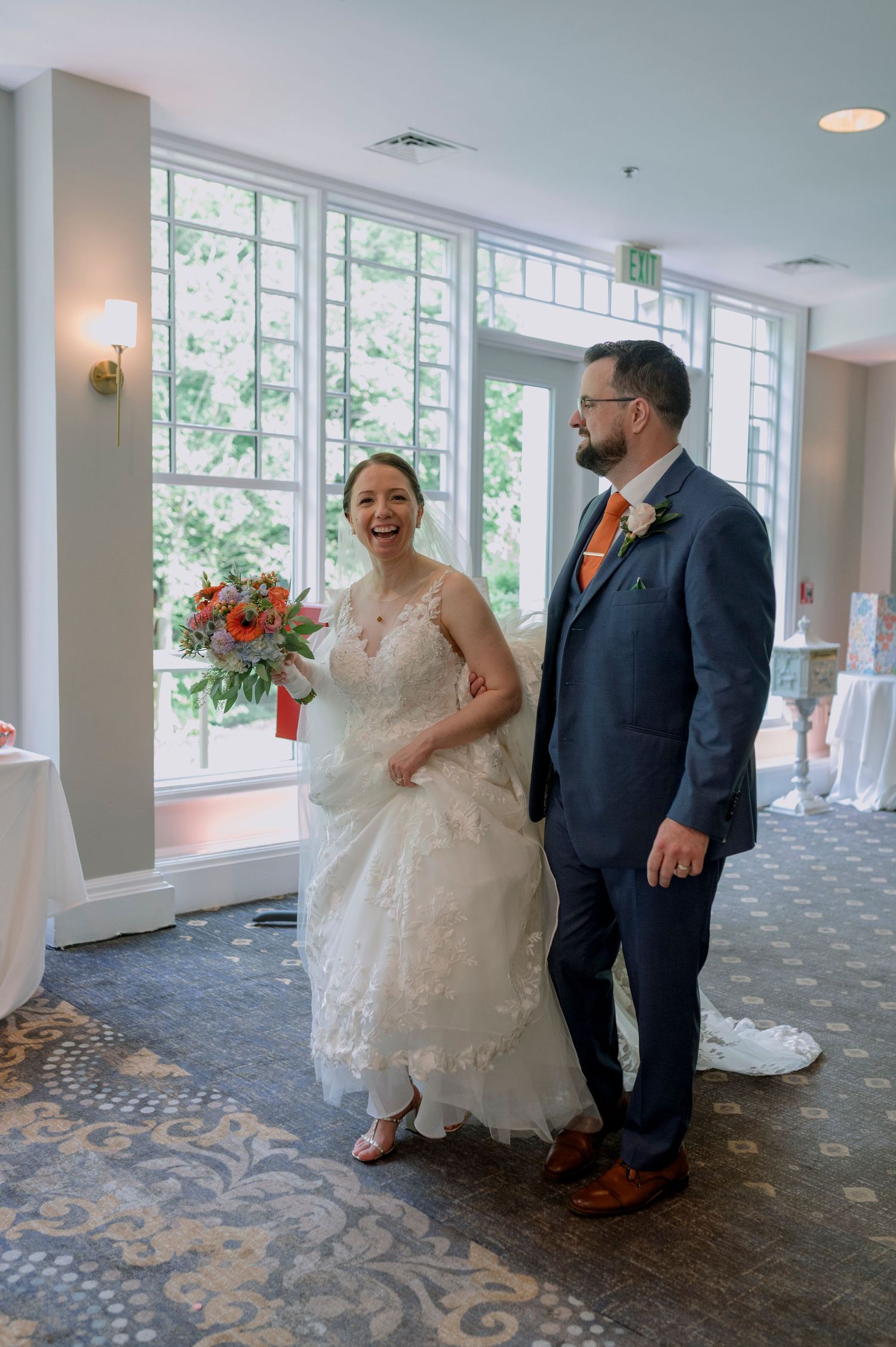 Joyful bride in lace gown holding orange bouquet stands with groom in navy suit at elegant wedding venue.