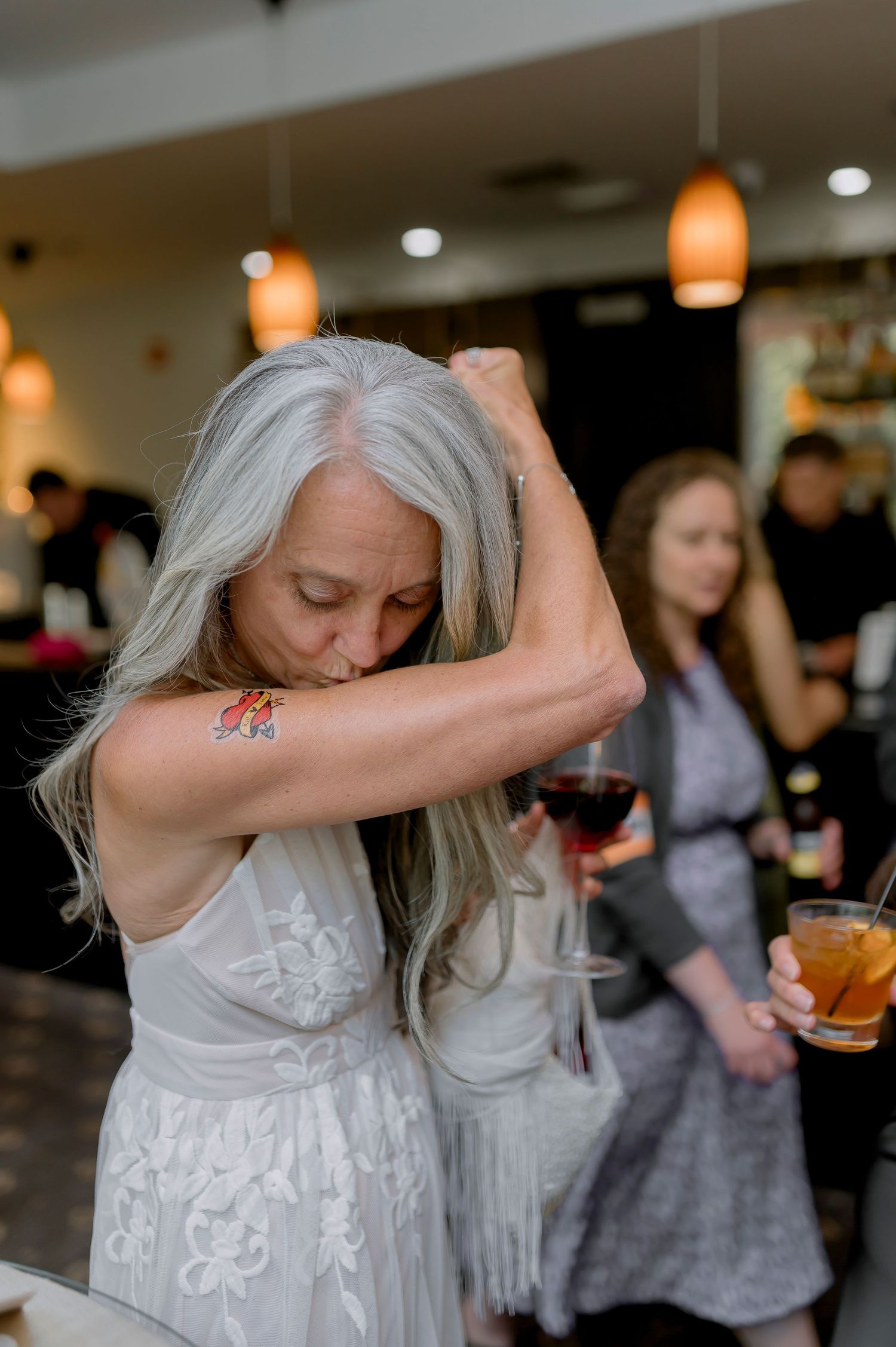 Older woman with long gray hair in white dress showing colorful tattoo on arm at indoor event.