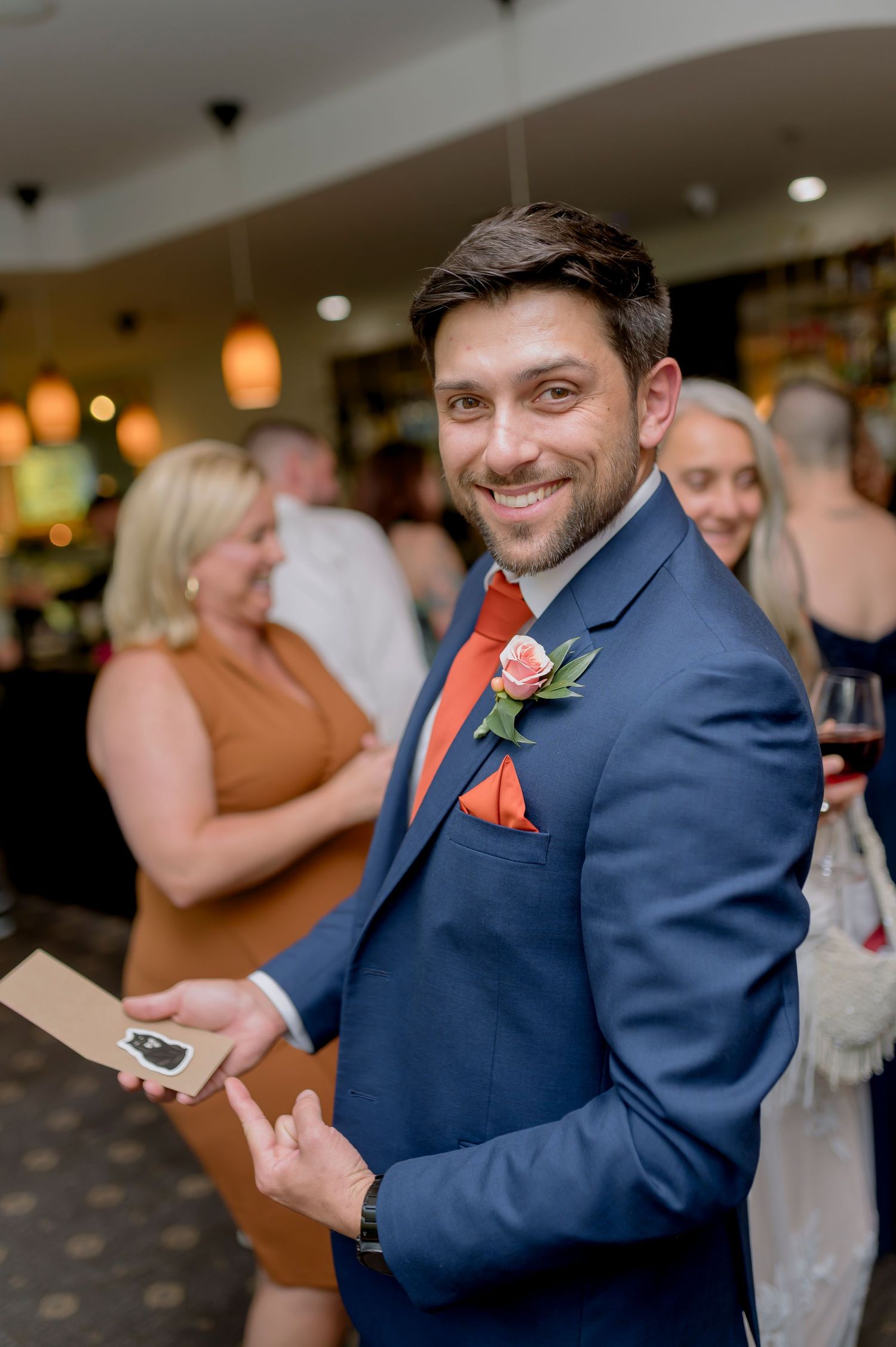 Smiling groom in navy blue suit with coral tie and boutonniere holds notes at elegant wedding reception.