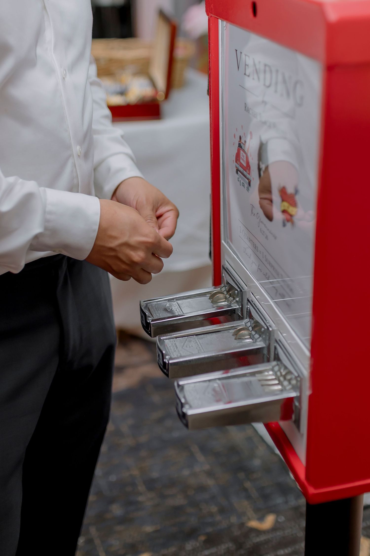 Man in white shirt retrieving items from a red vending machine with metal trays at an event.