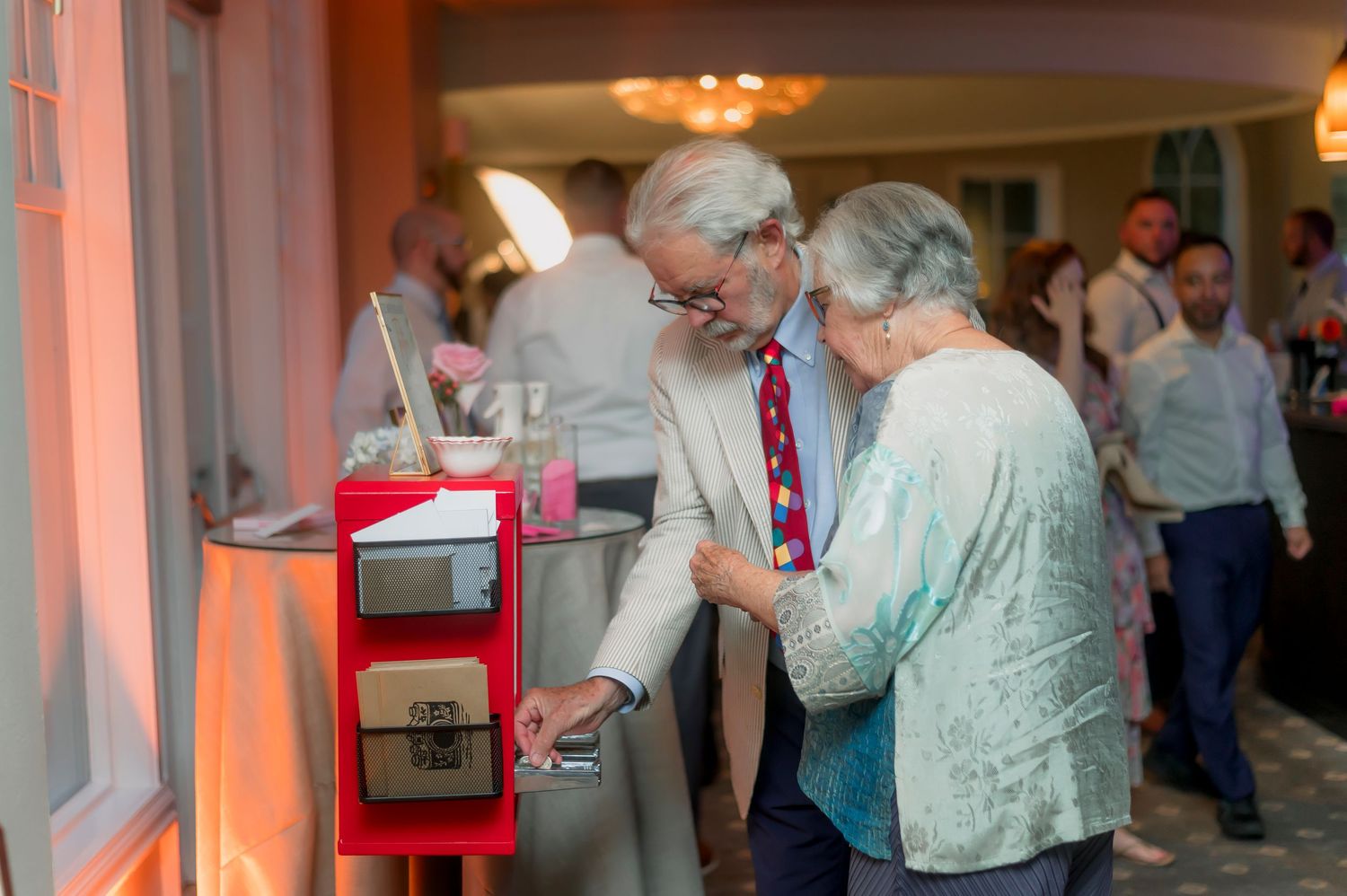 Elderly couple using a vintage red gumball machine at an elegant indoor social event.